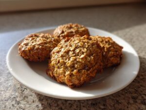 Close-up of four golden brown banana oatmeal cookies stacked slightly on a white plate, catching sunlight.