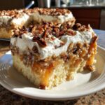 A moist slice of butter pecan praline poke cake topped with whipped cream and pecans, drizzled with caramel sauce.