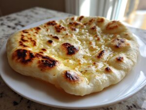 A single piece of freshly made naan bread, brushed with melted butter and showing charred bubbles, resting on a white plate.