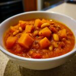 Close-up of a white bowl filled with rich, orange butternut squash chili featuring visible chunks of squash and white beans.