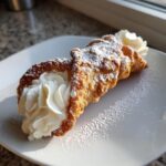 A single, freshly made cannoli recipe pastry filled with white cream and dusted generously with powdered sugar.