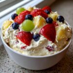 Close-up of a white bowl filled with creamy cheesecake fruit salad topped with strawberries, blueberries, grapes, and pineapple.