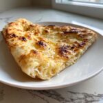 A close-up of a freshly baked slice of cheesy naan bread, golden brown and slightly charred on top, served on a white plate.