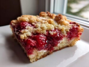 A close-up of a square slice of moist cherry dump cake with a crumbly topping on a white plate.