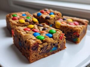 Four chewy M&M christmas cookie bars with colorful candies on a white plate.