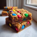A stack of two chewy M&M christmas cookie bars topped with colorful candies, showing a rich, dense center.