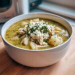 A close-up of a white bowl filled with rich, green chicken poblano soup recipe, topped with shredded chicken, white cheese, and cilantro.