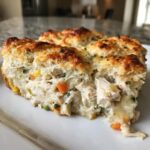 Close-up of a serving of chicken pot pie casserole showing creamy filling, chicken chunks, vegetables, and a golden biscuit topping.