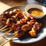 Close-up of perfectly grilled chicken satay skewers served on a grey plate next to a bowl of rich peanut sauce.