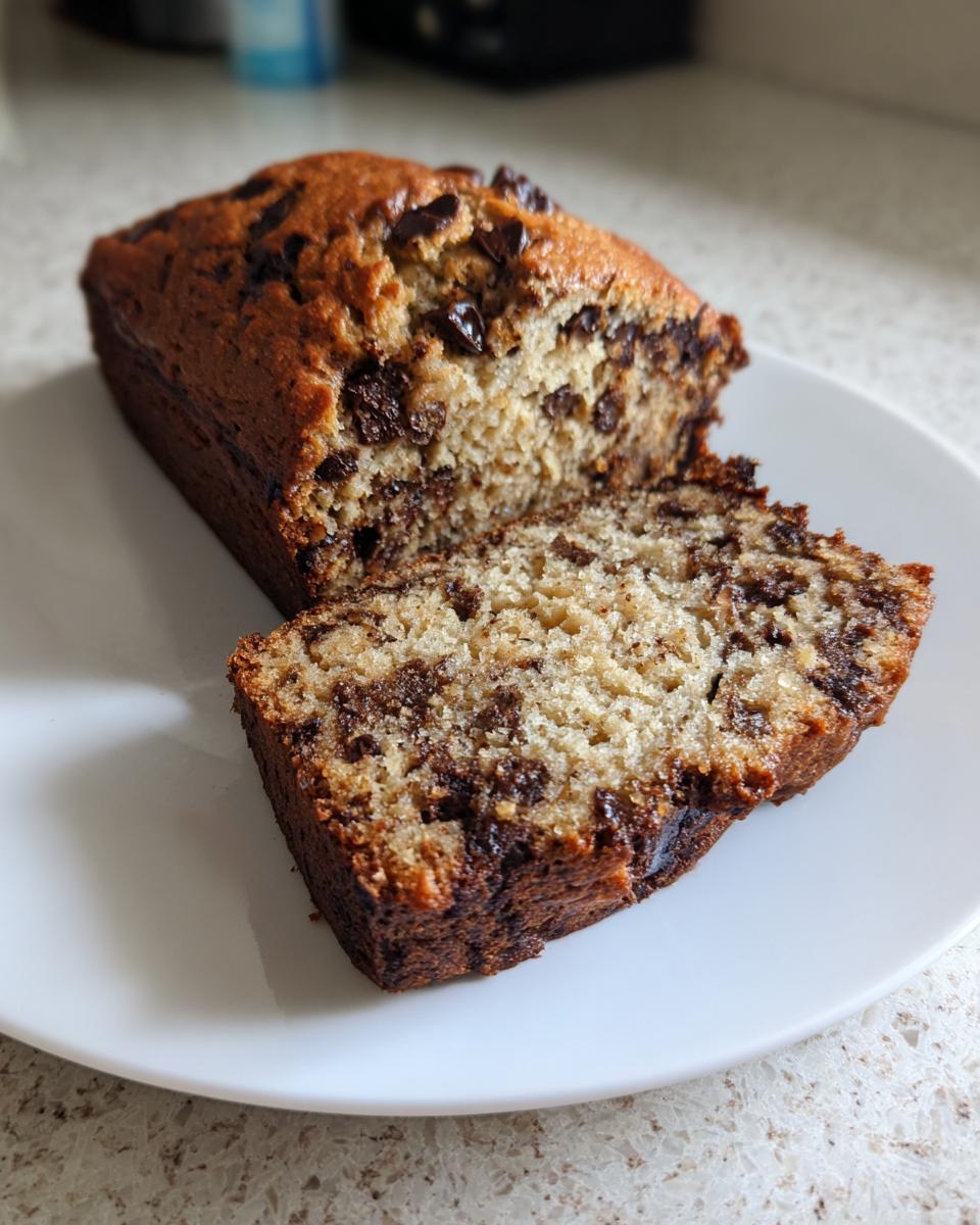 A loaf of moist chocolate chip banana bread with one slice cut and resting beside it on a white plate.