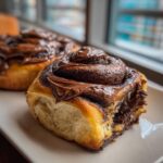 A close-up of a fluffy, freshly baked chocolate cinnamon rolls topped with melted chocolate glaze.
