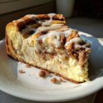 A close-up shot of a thick slice of cinnamon roll cheesecake drizzled with white icing on a white plate.