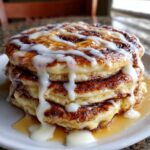 A tall stack of fluffy cinnamon roll pancakes drizzled heavily with white cream cheese icing.
