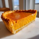 A close-up, sunlit slice of classic pumpkin pie showing the rich orange filling and flaky crust.