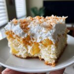 A moist slice of coconut poke cake topped with whipped cream and toasted coconut flakes.