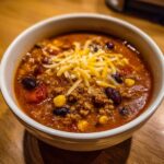 Close-up of a bowl of rich cowboy soup filled with ground meat, beans, corn, and topped with shredded cheddar cheese.