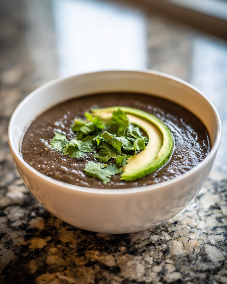 A close-up of a bowl of thick, dark black bean soup topped with fresh cilantro and sliced avocado.