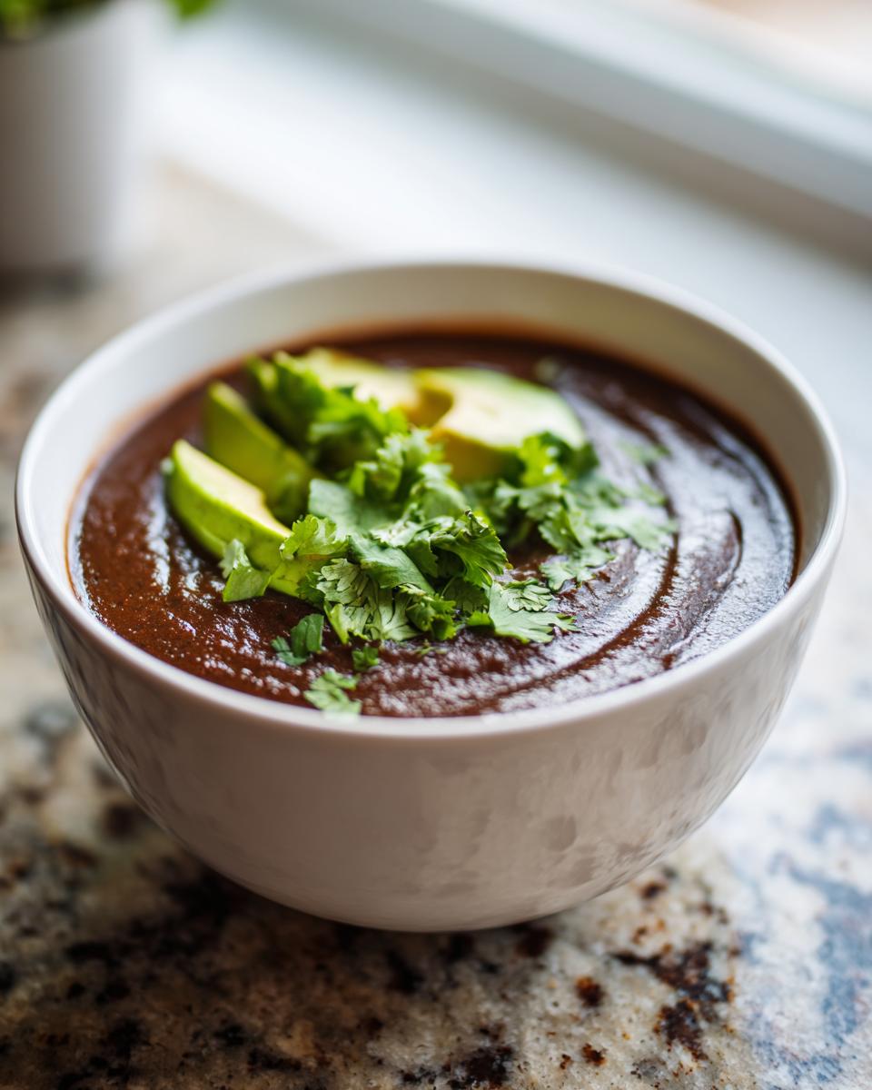 A close-up of a bowl of rich, dark black bean soup topped with sliced avocado and fresh cilantro.