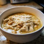 Close-up of a white bowl filled with creamy chicken chili, featuring shredded chicken, white beans, and topped with melted cheese.