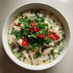 Overhead shot of a white bowl filled with creamy chicken coconut soup, topped with fresh cilantro, sliced red chilies, and mushrooms.