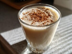 A close-up of a glass filled with creamy coquito, topped with a dusting of ground cinnamon.