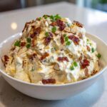 A large white bowl filled with creamy Neiman Marcus dip, topped generously with bacon bits, sliced almonds, and green onions.