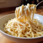 Close-up of spaghetti coated in rich homemade alfredo sauce, lifted by a fork, topped with grated cheese.