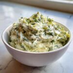 A white bowl filled high with chunky, creamy pickle dip, garnished generously with fresh dill.