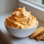 A white bowl filled with fluffy, orange pumpkin fluff dip, served next to a stack of square crackers.