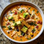 Close-up of a bowl of creamy tortilla soup, loaded with shredded chicken, corn, black beans, and topped with avocado cubes and tortilla strips.