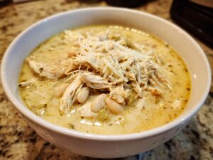 A close-up of a white bowl filled with creamy white chicken chili, featuring shredded chicken, white beans, and topped with shredded cheese.