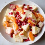 Close-up of a creamy winter fruit salad featuring diced apples, mandarin segments, and bright pomegranate seeds.