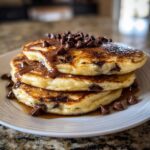 A stack of three fluffy chocolate chip pancakes drizzled with chocolate syrup and topped with chocolate chips.