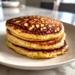 A stack of three fluffy coconut flour pancakes with golden-brown, bubbly tops served on a white plate.