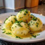 Four fluffy potato dumplings served on a white plate, glistening with melted butter and topped with fresh chopped parsley.