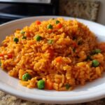 A close-up of fluffy, orange-colored spanish rice mixed with bright green peas and diced carrots on a white plate.