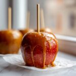 Close-up of a shiny red caramel apple with thick caramel dripping down the sides, set against a blurred background.