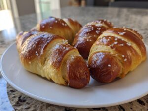 Four freshly baked pretzel dogs with shiny, dark brown crust and coarse salt, arranged on a white plate.