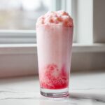 A tall glass filled with frothy pink strawberry milk, topped with a mound of foam.