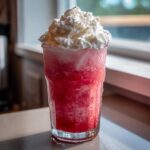 A tall glass of icy, bright red frozen pink lemonade topped high with whipped cream.