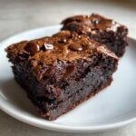 Close-up of a fudgy date brownies square on a white plate, showing a shiny, crackly top and melted chocolate chips.