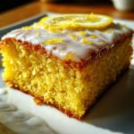A close-up, vibrant slice of fudgy lemon brownies topped with white glaze, zest, and a lemon slice.