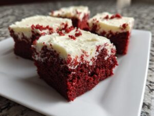 Four squares of fudgy red velvet brownies topped with thick cream cheese frosting and red crumbs.
