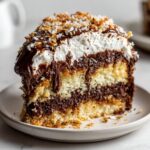 A decadent slice of german chocolate poke cake showing layers of light cake, dark chocolate filling, whipped topping, and toasted coconut.