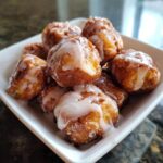 A close-up of several golden brown cinnamon roll bites piled in a white square bowl, drizzled with thick white icing.