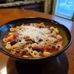 A close-up of a hearty bowl of pasta e fagioli, featuring pasta, beans, ground meat, and topped with grated Parmesan cheese.