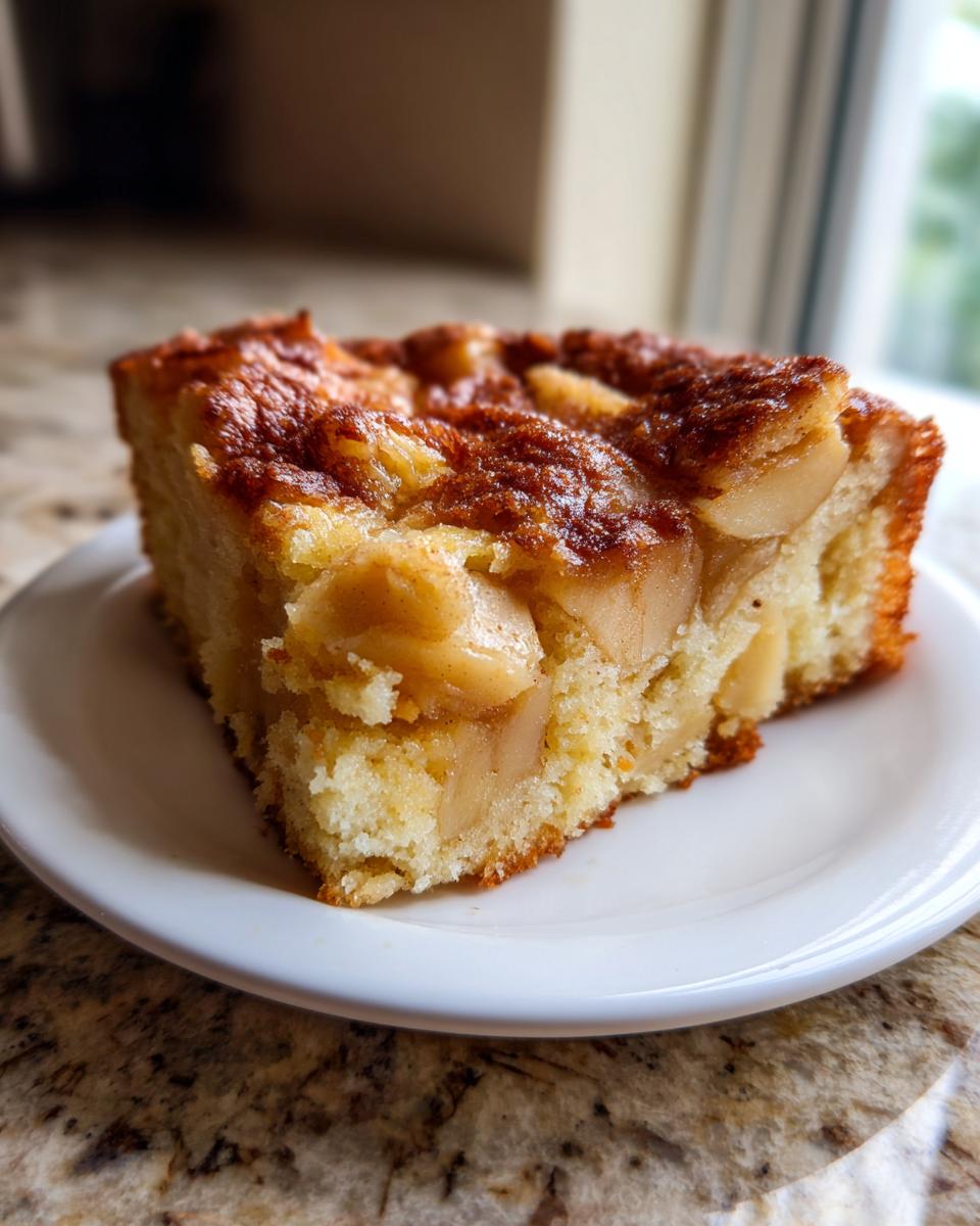 A close-up of a single slice of jewish apple cake on a white plate, showing moist crumb and caramelized apple topping.