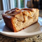A close-up of a moist slice of jewish apple cake with visible chunks of baked apple on a white plate.