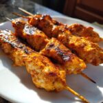 Four grilled bang bang chicken skewers resting on a white plate, showing char marks and a glossy orange coating.
