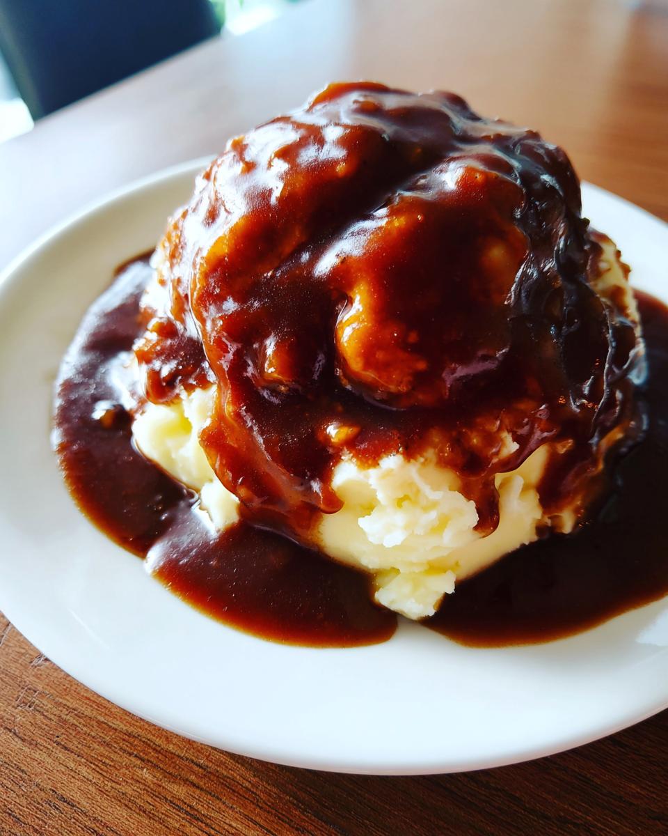 A mound of mashed potatoes smothered in rich, dark brown chicken gravy on a white plate.